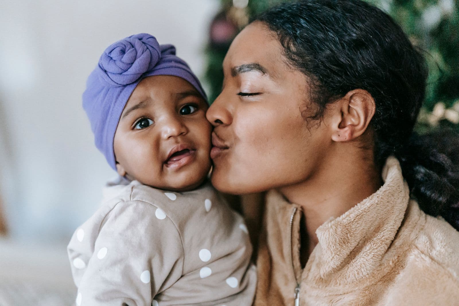 Mãe beijando a bochecha do bebê, que usa um turbante lilás e olha para a câmera.