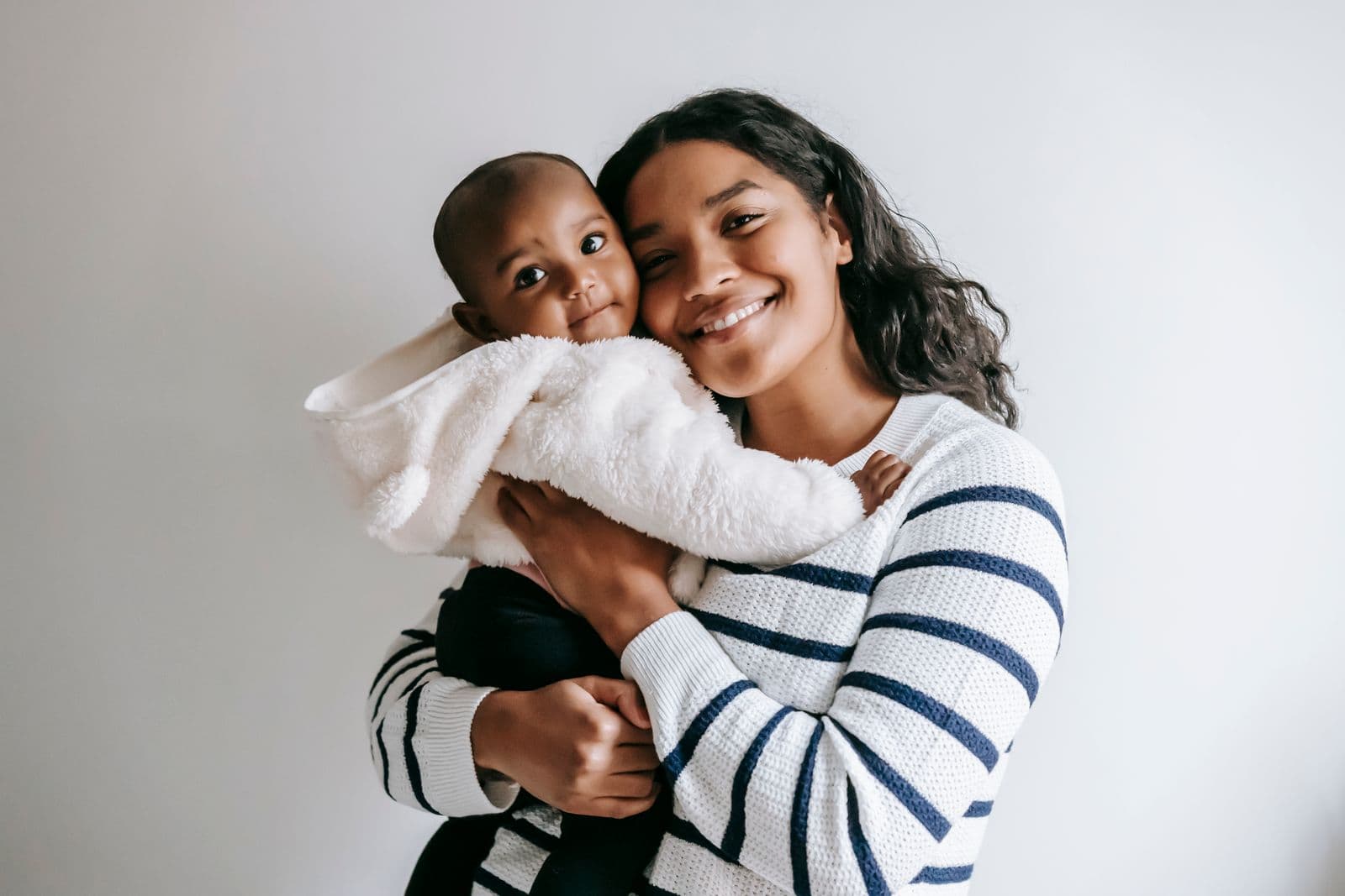 Mãe de suéter listrado sorrindo para a câmera com o bebê no colo, enrolado em uma manta branca felpuda.
