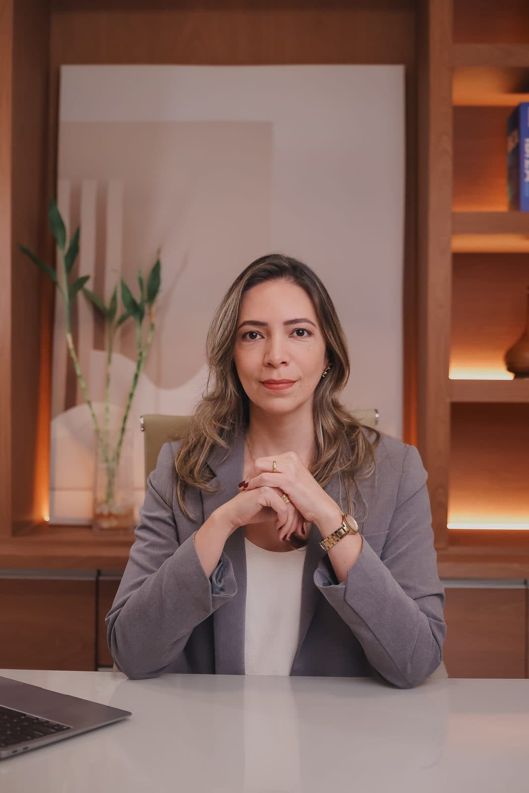 Dra. Sarah Mesquita à mesa do escritório, de blazer cinza, com as mãos entrelaçadas.
