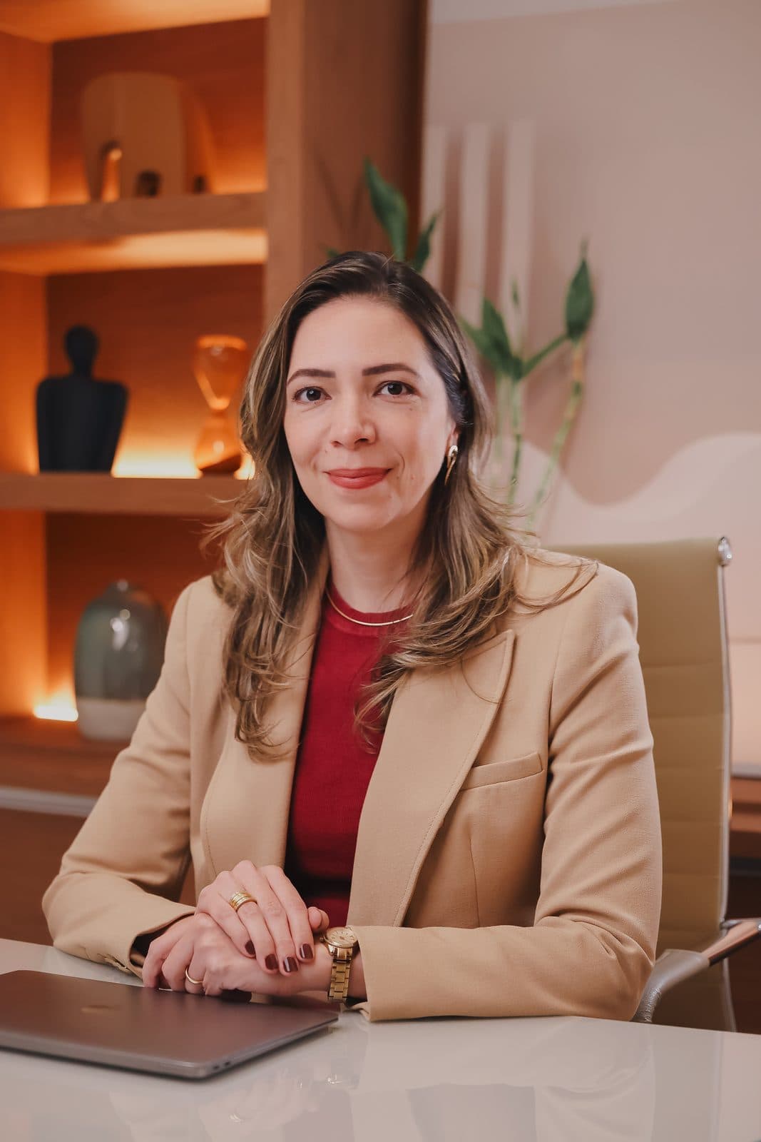 Dra. Sarah Mesquita sentada à mesa do escritório, de blazer bege, com as mãos apoiadas sobre o notebook fechado.